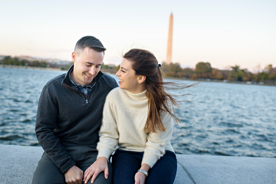 A fun engaged couple sitting along the tidal basin with the washington monument behind them.
